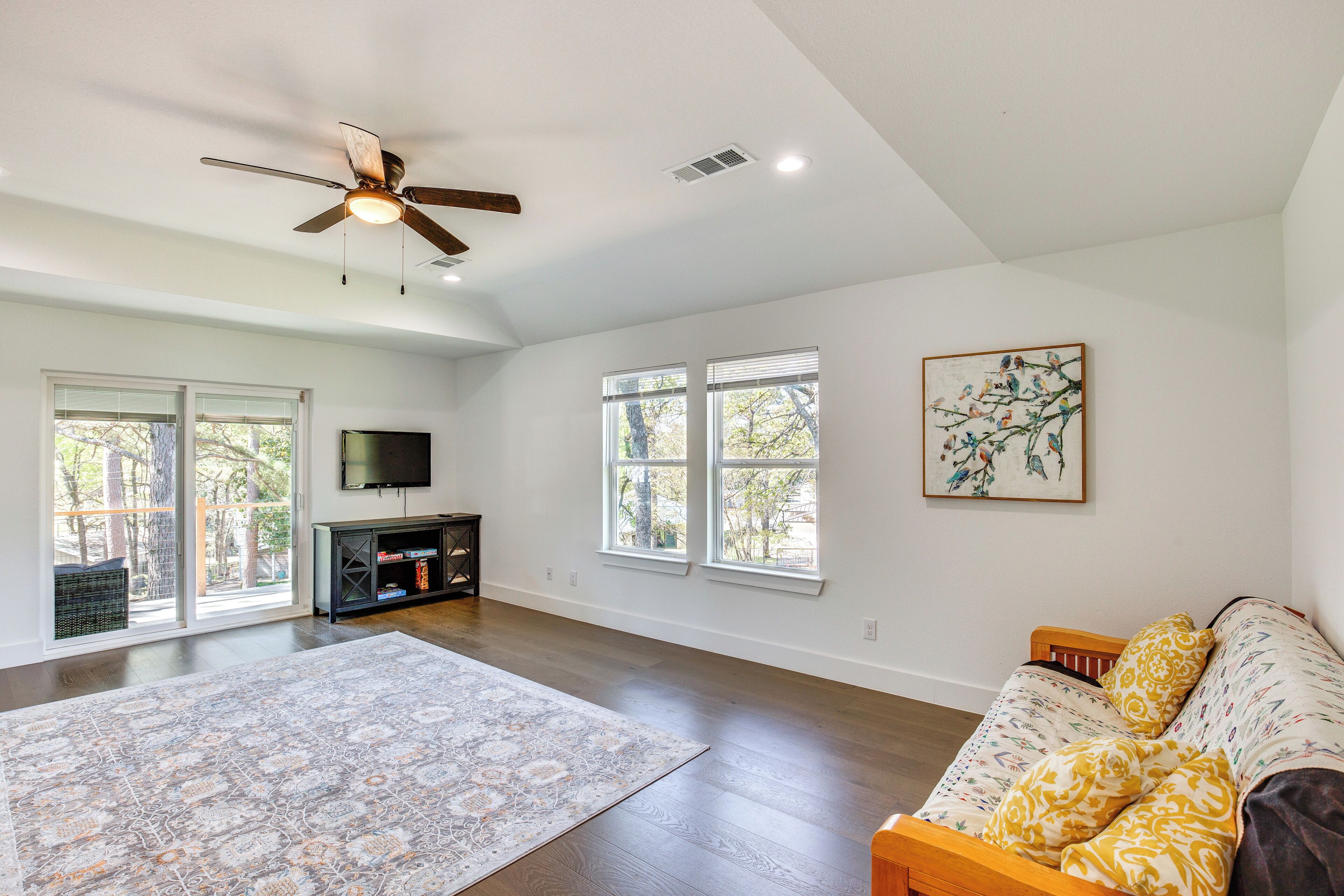 A spacious living room with hardwood floors, a ceiling fan, a large window, a flat-screen TV, and a sofa with patterned cushions.