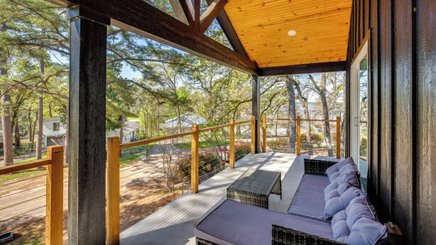 A covered outdoor seating area with a ceiling fan, wooden beams, and a view of trees and a road. - Fire Pit + Grill: Lakeside Gun Barrel City Home! (Mabank)