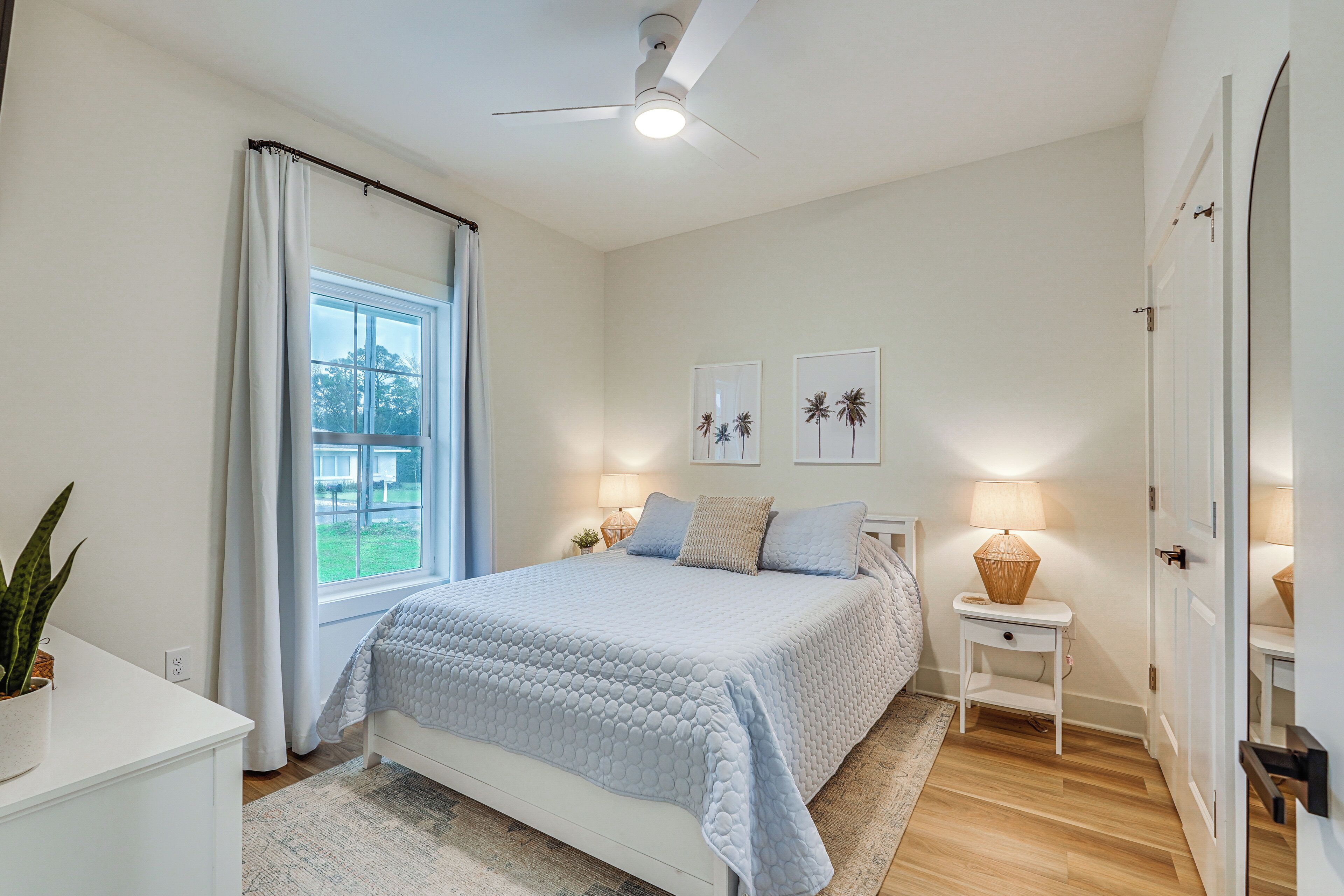 Un dormitorio con cama, una mesita de noche con lámpara, una ventana con cortinas y un ventilador de techo.