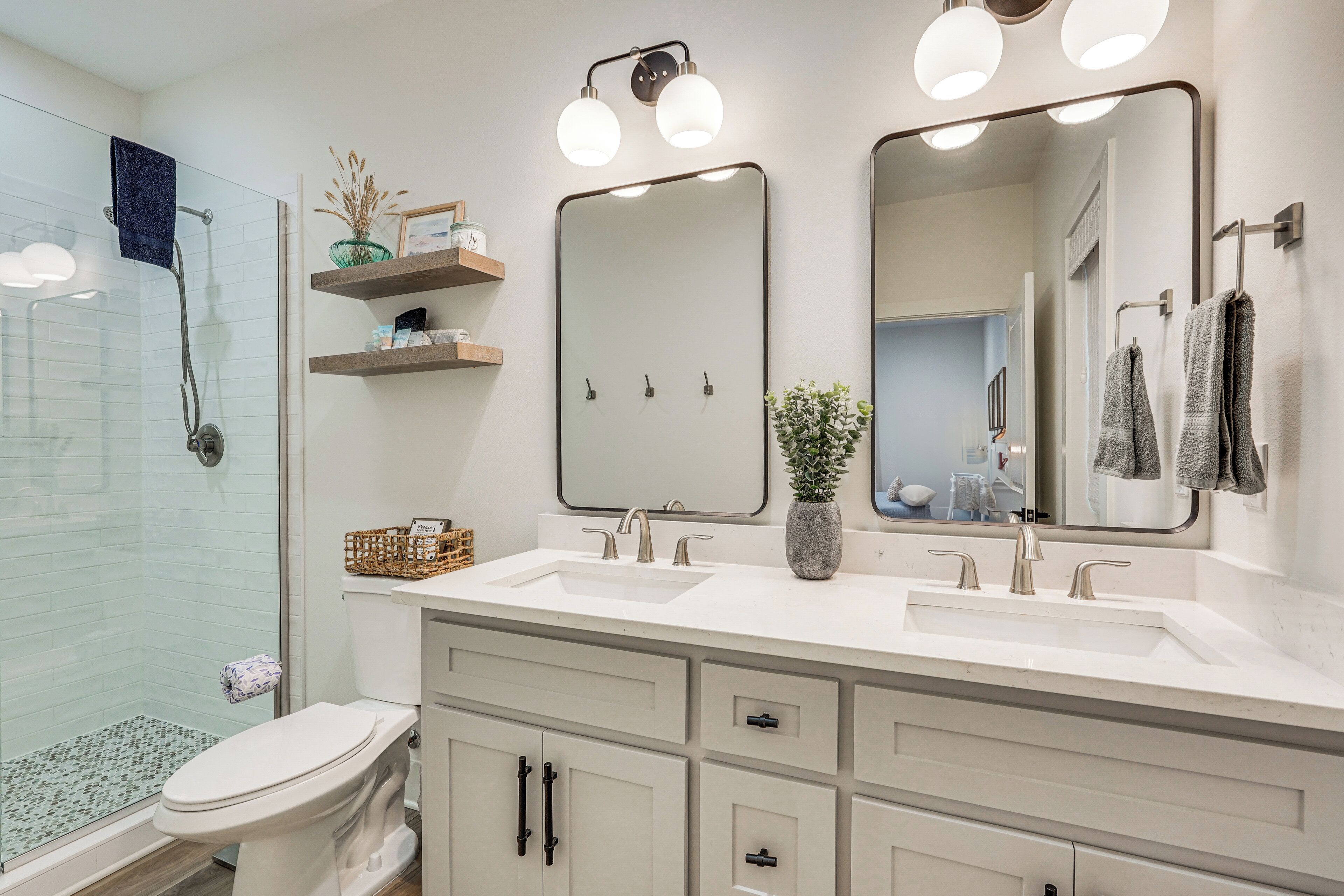 A modern bathroom with a glass shower enclosure, a white sink with two mirrors, and a toilet with a basket of toiletries.