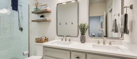 A modern bathroom with a glass shower enclosure, a white sink with two mirrors, and a toilet with a basket of toiletries.
