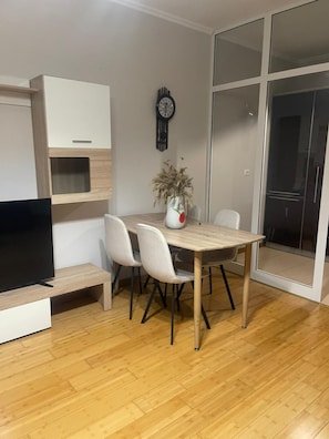 A modern living room with a flat-screen TV, a wooden dining table, white chairs, a potted plant, and a sliding glass door leading to a balcony. - Kalpazo Home Skanderbeg Square (Tirana)