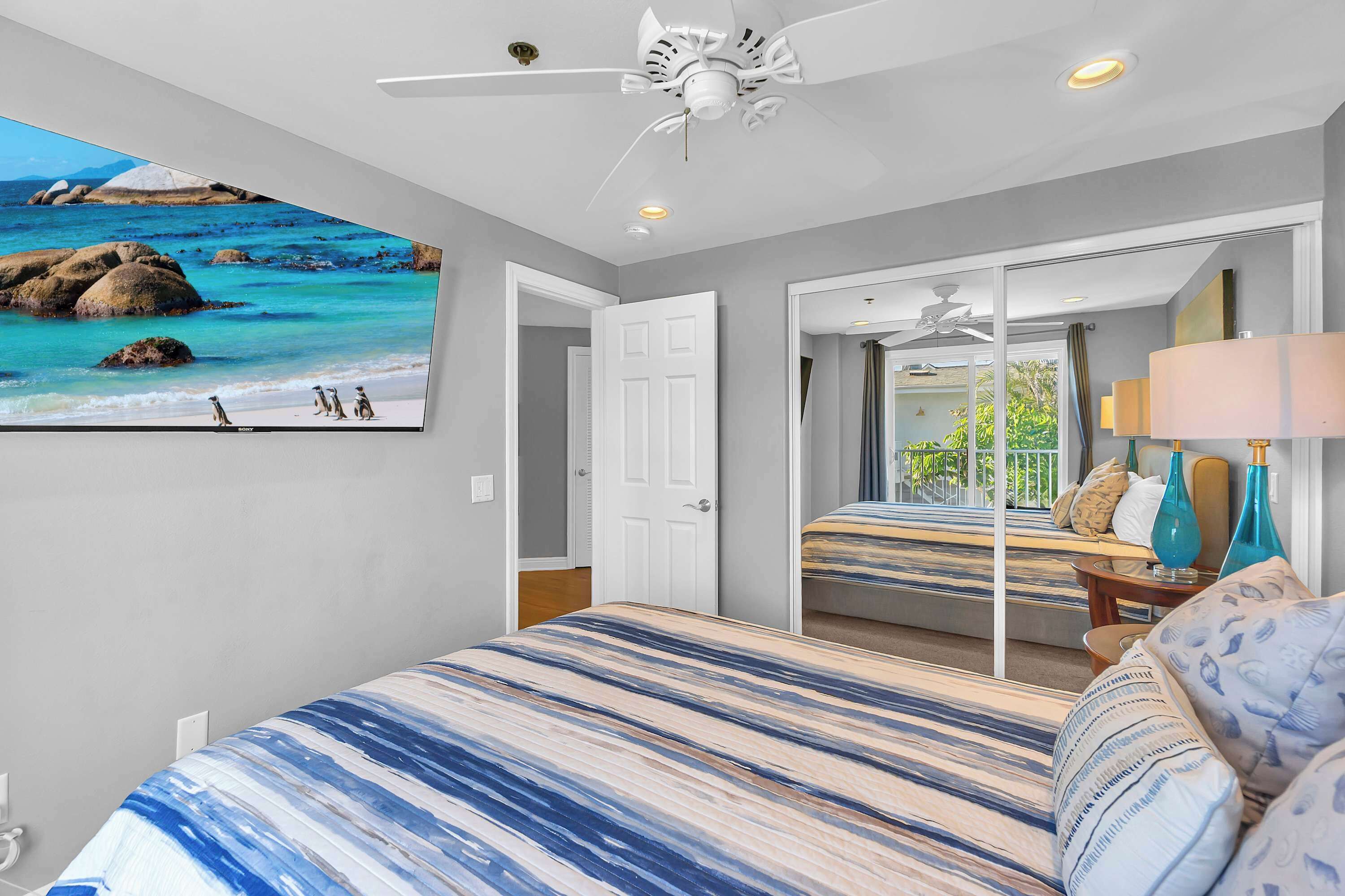 Une chambre avec un grand lit, un ventilateur de plafond, un téléviseur diffusant une scène de plage et une porte donnant sur une autre pièce.