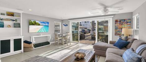 A spacious living room with a flat-screen TV, a wooden coffee table, a beige sofa, and a large sliding glass door leading to an outdoor area.