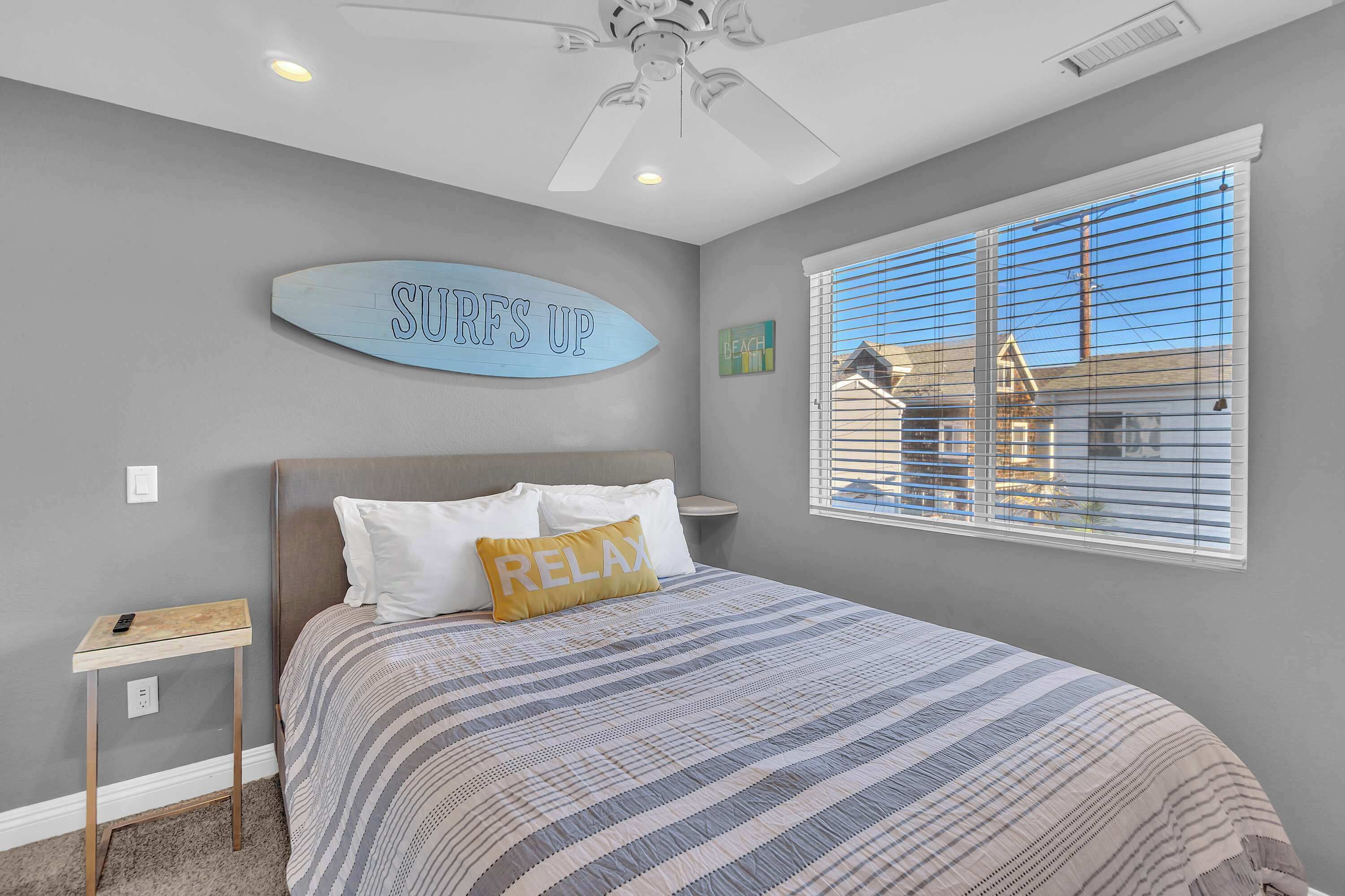 A bedroom with a bed, a ceiling fan, a window with blinds, and a sign on the wall.
