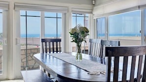 A dining area with a wooden table and chairs, a vase with flowers, and large windows offering a view of the ocean.
