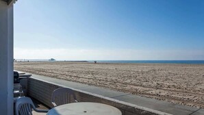 Une vue sur la plage depuis un balcon, avec une table et des chaises, une barrière en béton et une jetée au loin.