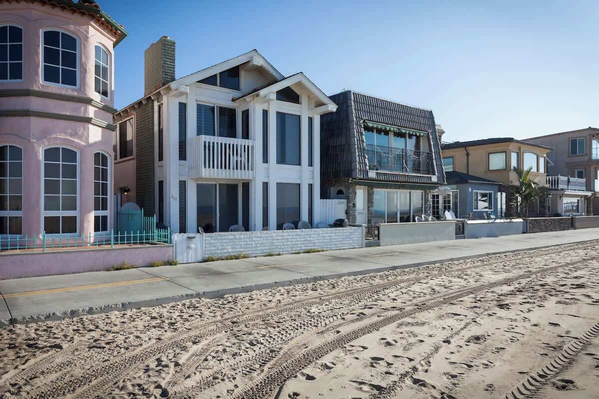 Une propriété située en bord de mer, comprenant une grande maison dotée d’un balcon, une dépendance plus petite attenante et une plage de sable en premier plan.