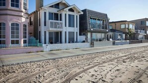 Une propriété située en bord de mer, comprenant une grande maison dotée d’un balcon, une dépendance plus petite attenante et une plage de sable en premier plan.