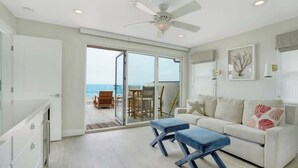 Uma sala de estar espaçosa com uma porta de vidro deslizante que dá para uma varanda com vista para o oceano. Há um sofá branco, dois pufes azuis e um ventilador de teto.