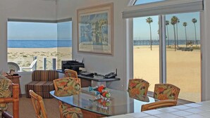 Una habitación amplia con una gran mesa de vidrio, asientos cómodos, un escritorio con computadora y un cuadro. Un ventanal ofrece vista a la playa y al océano.