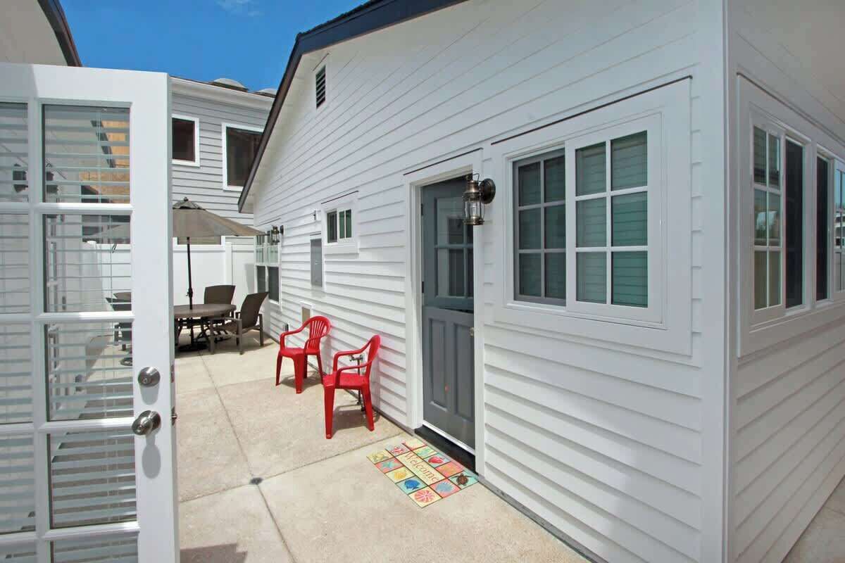 A white house with a gray door, a patio with a table and chairs, and a colorful mat.