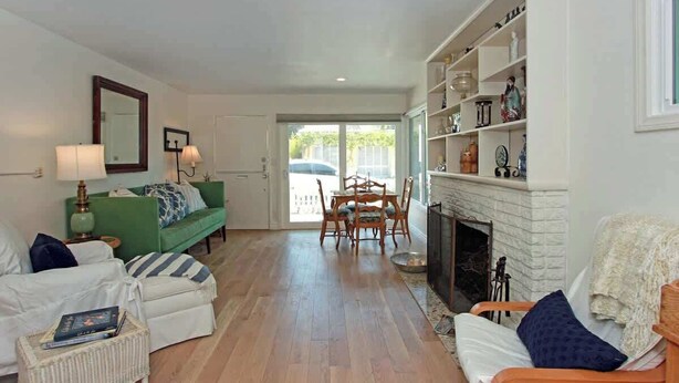 A living room with a green sofa, white armchairs, a dining table with chairs, a fireplace, and a large window. - 1009 W Bay | Quaint Cottage Steps to Sand (Newport Beach)