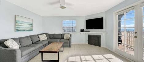 A living room with a grey sectional sofa, a wooden coffee table, a ceiling fan, a flat-screen TV, and a fireplace.