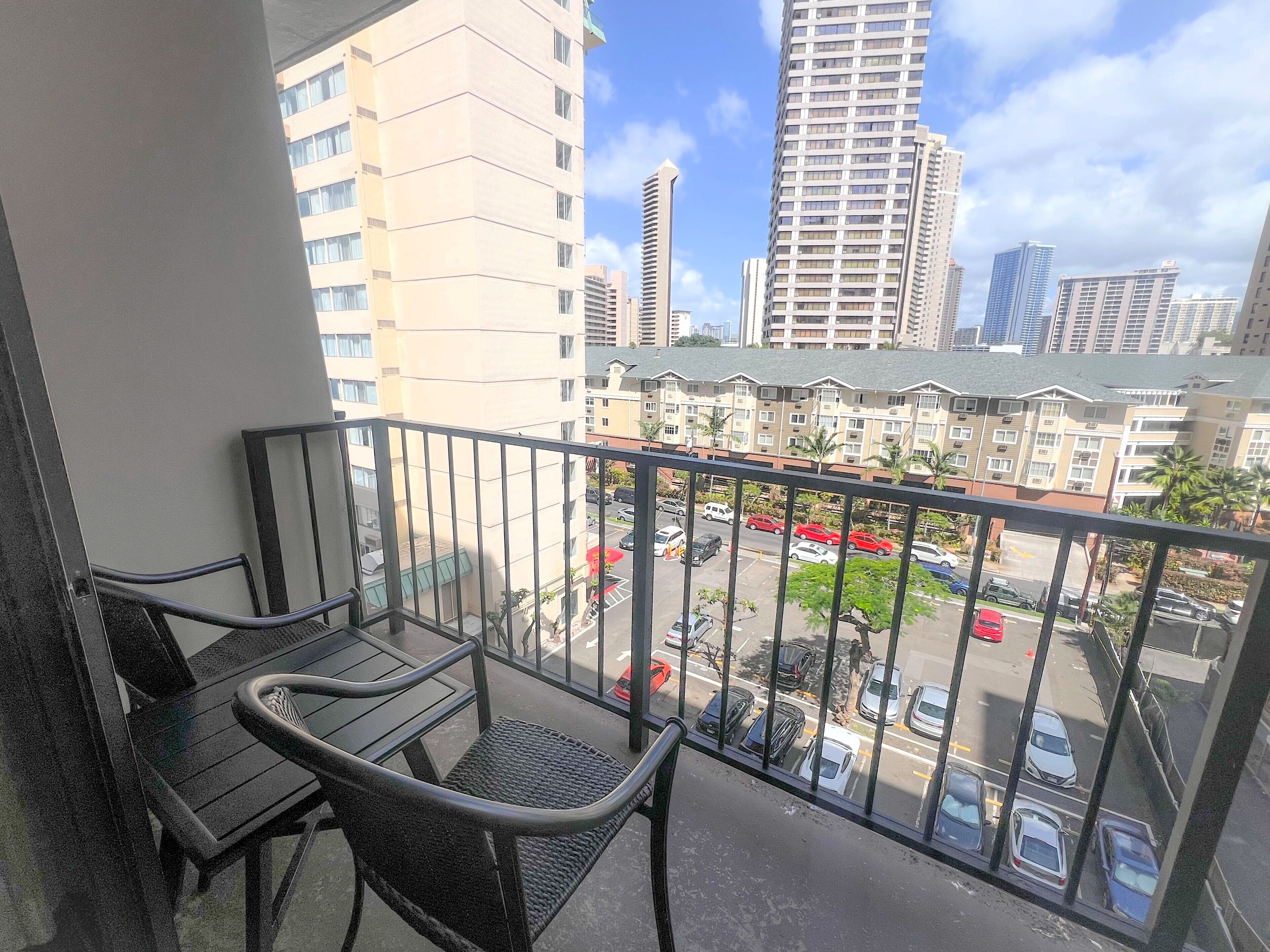 A balcony with two wicker chairs, overlooking a parking area and several buildings.