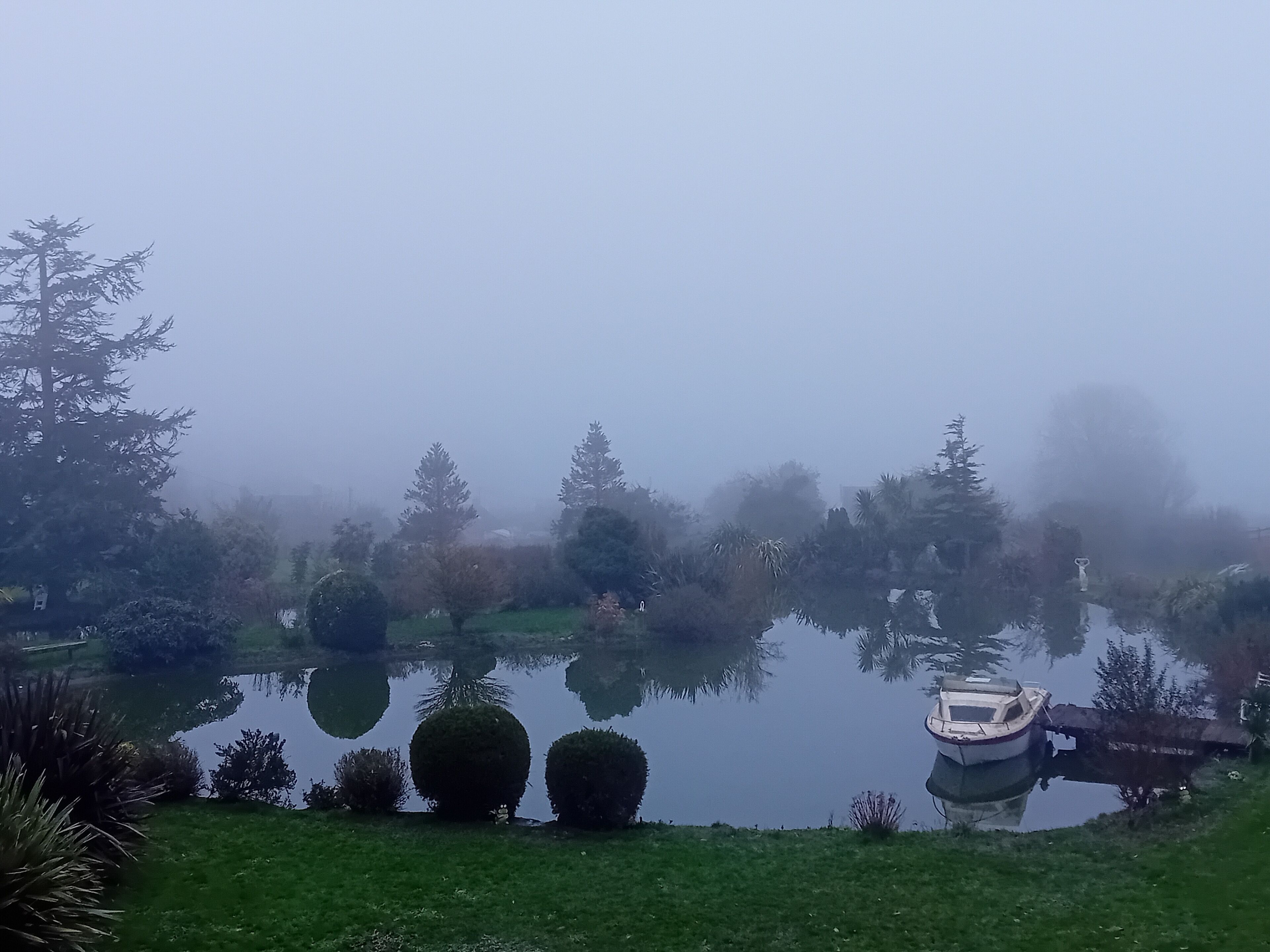 A calm lake with a boat docked on the right, surrounded by neatly trimmed bushes and trees.