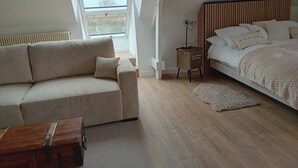 Et soveværelse med trægulv, seng med hvidt sengetøj, sofa med stribede puder, et lille træbord og en radiator.