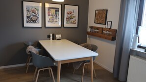 A dining area with a white table and four chairs, framed artwork on the wall, and a wooden shelf with kitchen items.