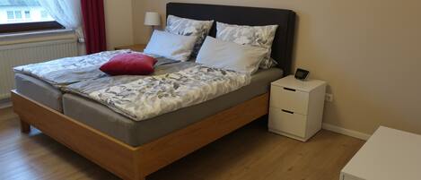 A neatly made bed with a floral bedspread, a red pillow, and a bedside table with a lamp and a small electronic device.