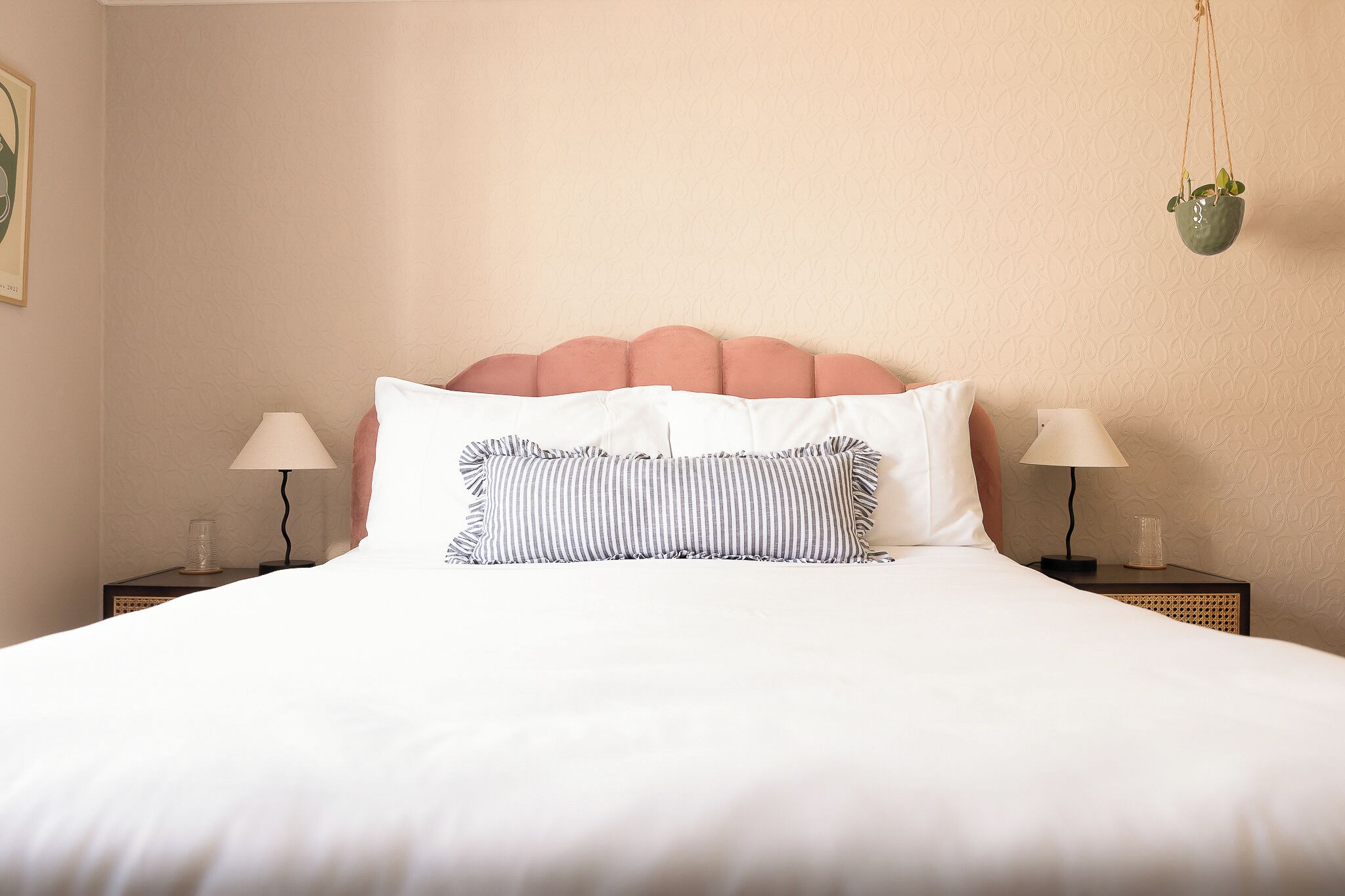 A neatly made bed with white bedding, two bedside tables with lamps, and a hanging plant.