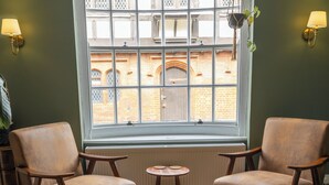 Quarto com duas poltronas de couro, uma mesa redonda pequena e uma janela com vista para um edifício de tijolos.