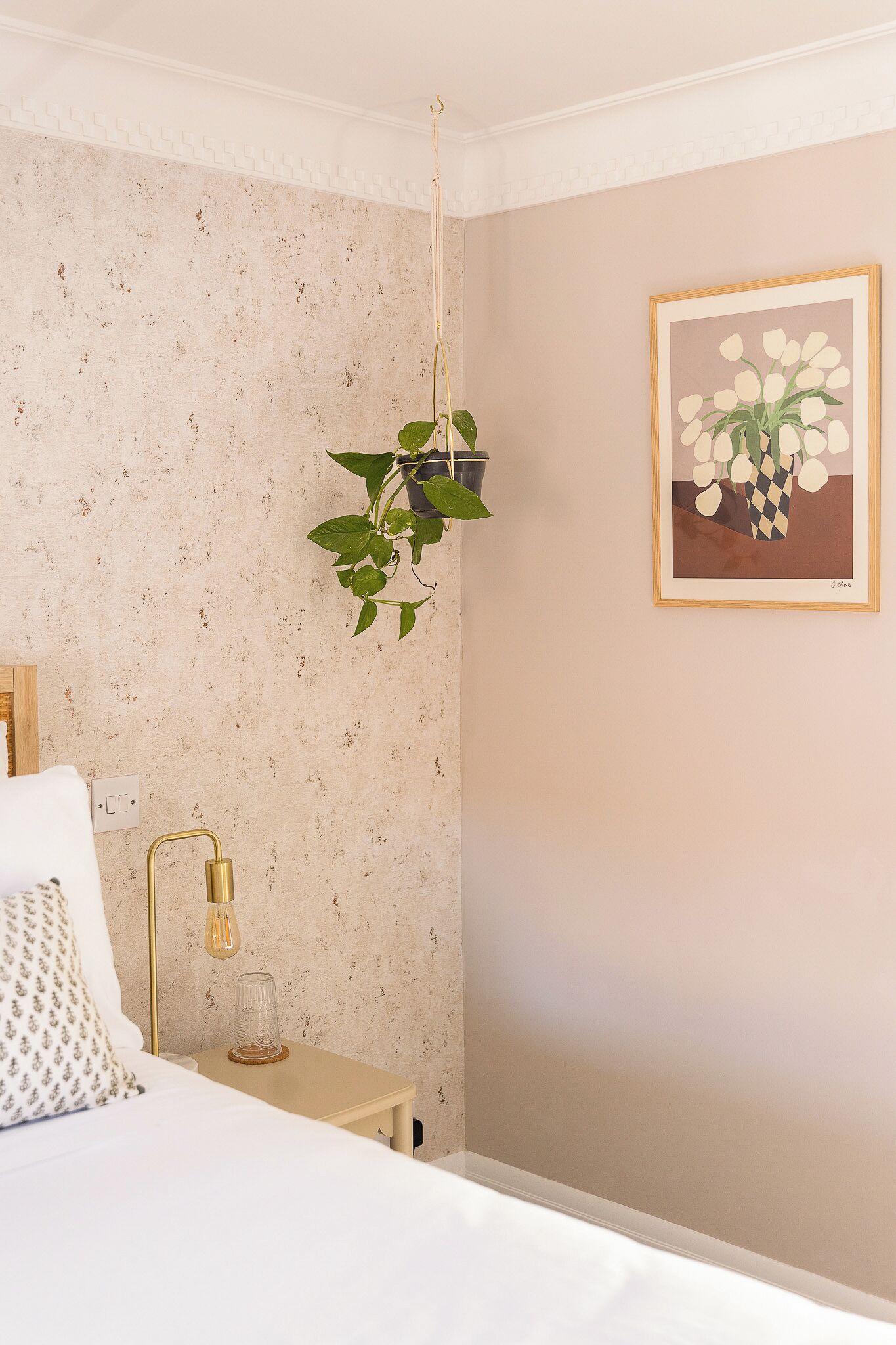 Un dormitorio con cama, mesita de noche, lámpara y una planta colgante.