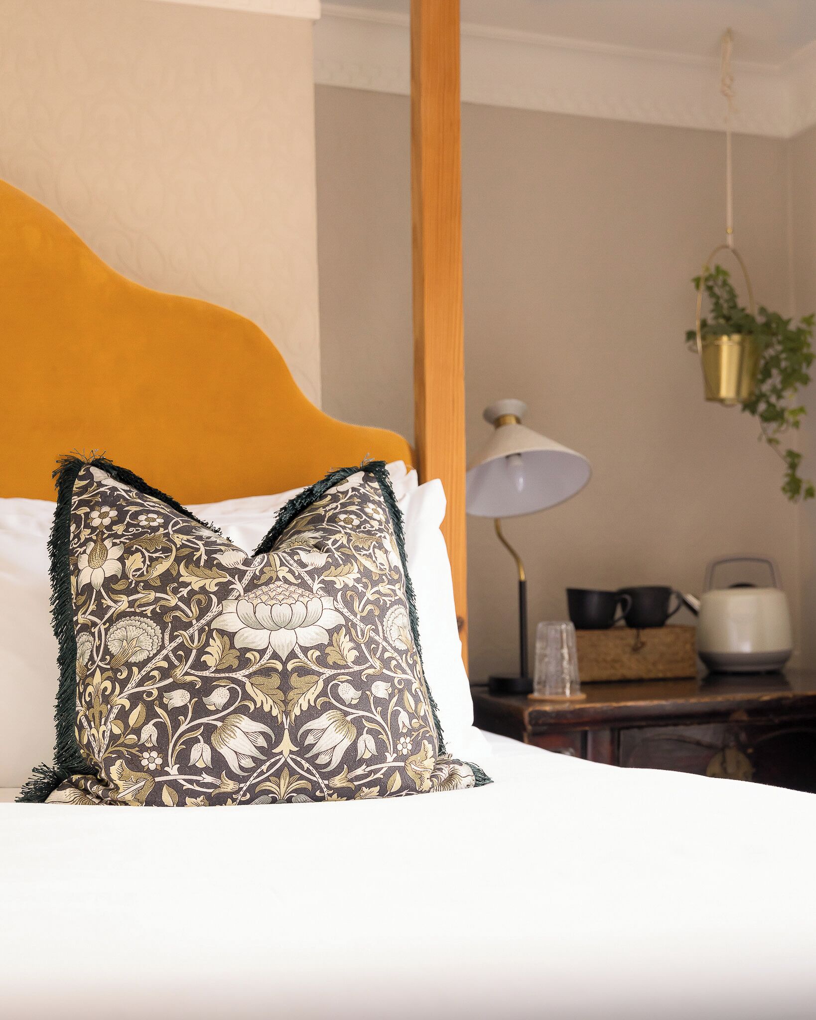 A bed with a patterned pillow, a wooden headboard, a bedside table with a lamp, and a hanging plant.