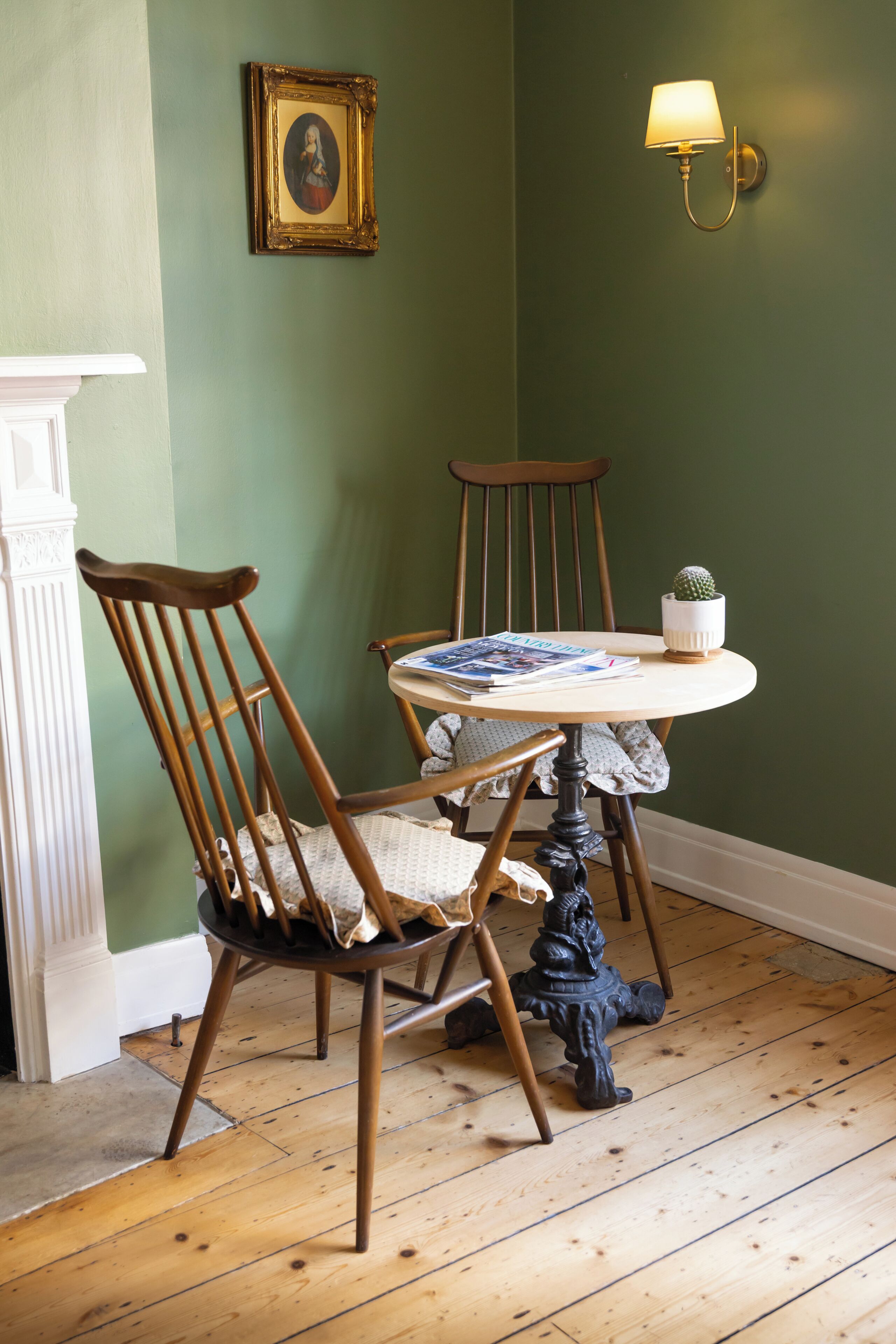 A dining area with a round table, two wooden chairs, a small potted plant, and a wall-mounted lamp.