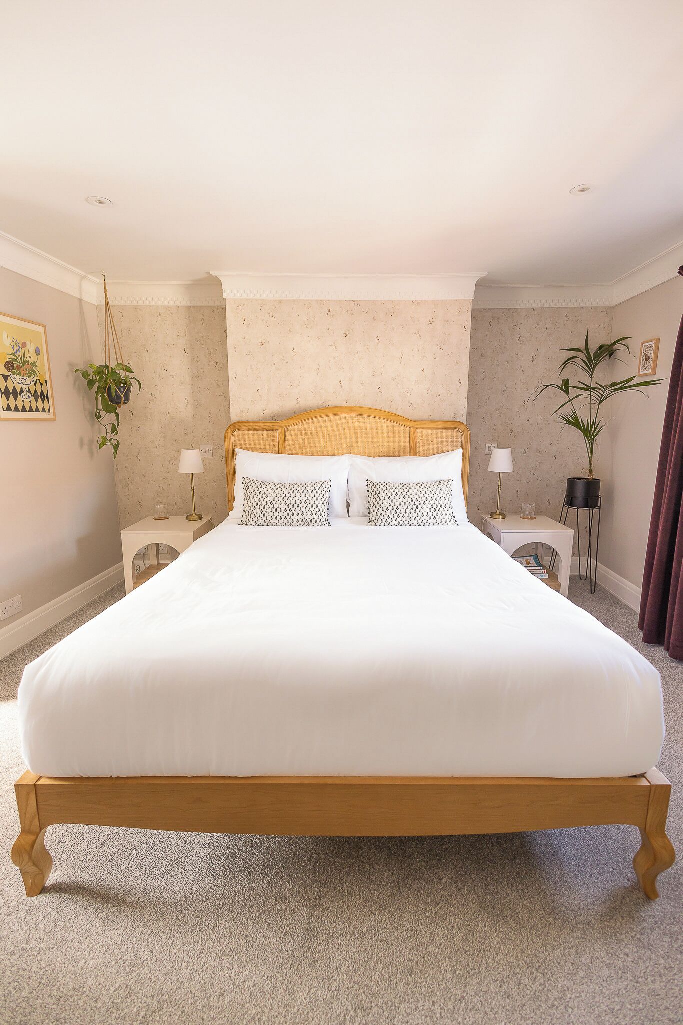 A neatly made bed with a wooden frame, two bedside tables with lamps, a potted plant, and a wall with a floral pattern.