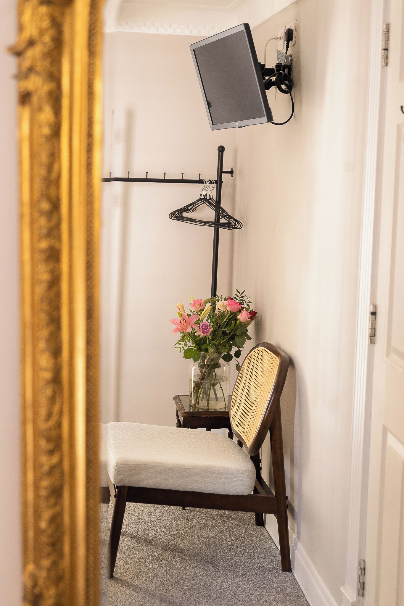 A room with a wall-mounted TV, a chair, a small table with a vase of flowers, and a coat rack.