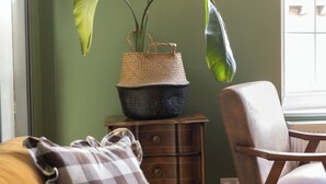 Um quarto com parede verde, mesa lateral de madeira com abajur, uma cadeira e um sofá com almofada xadrez.