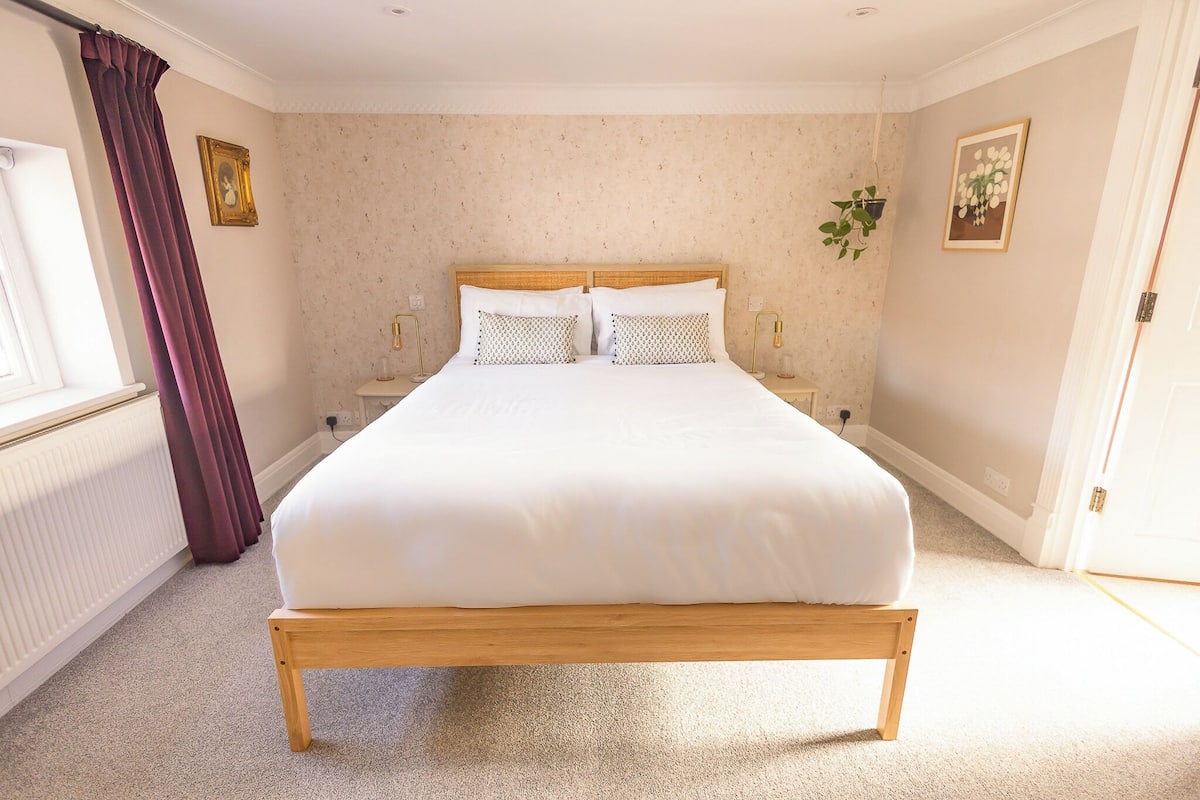 A neatly made bed in a room with a plain wall, a window with curtains, and a door.