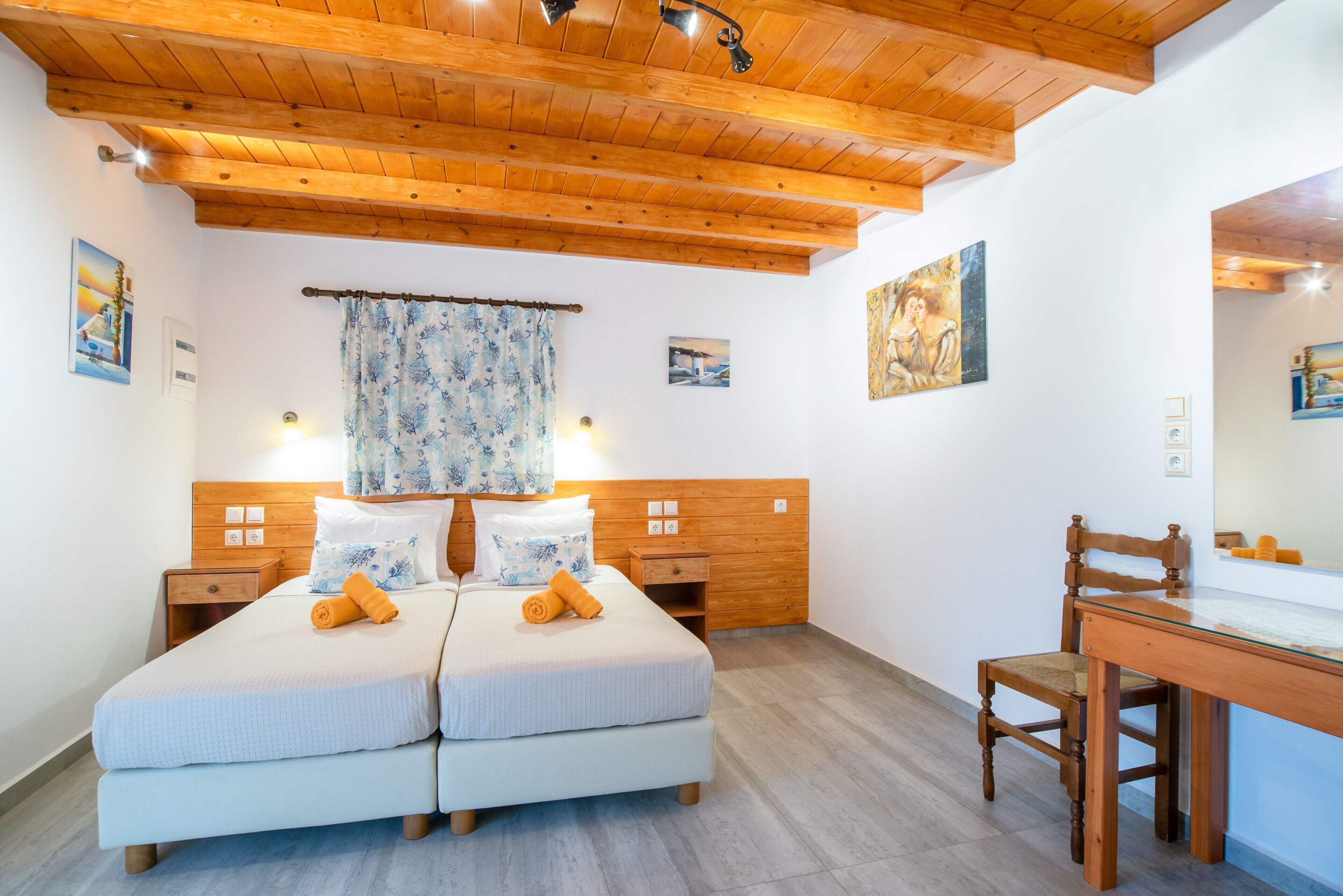 A bedroom with two beds, wooden headboard, a desk with a chair, and artwork on the walls.