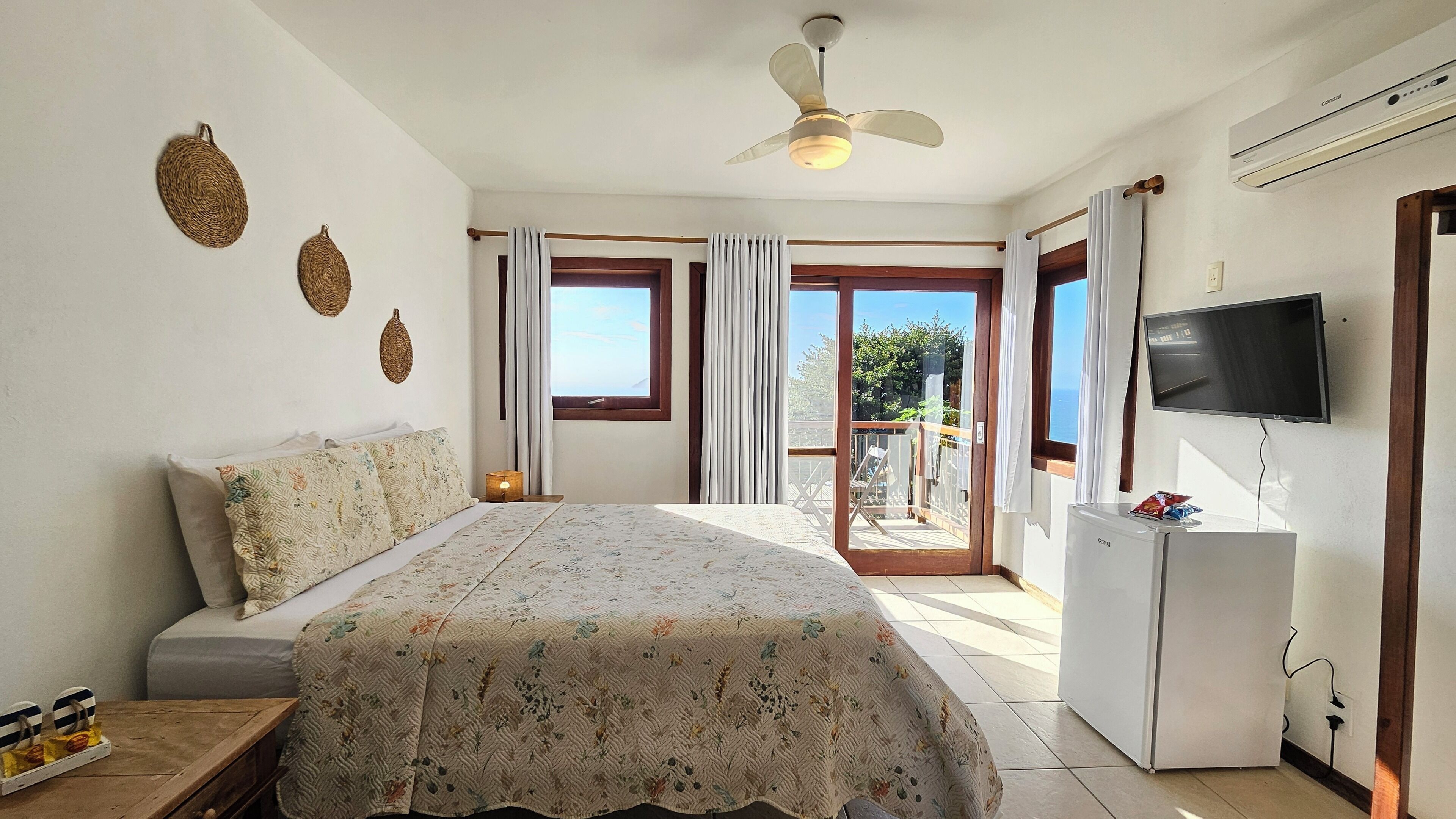A bedroom with a bed, a ceiling fan, a TV, and a balcony with curtains.