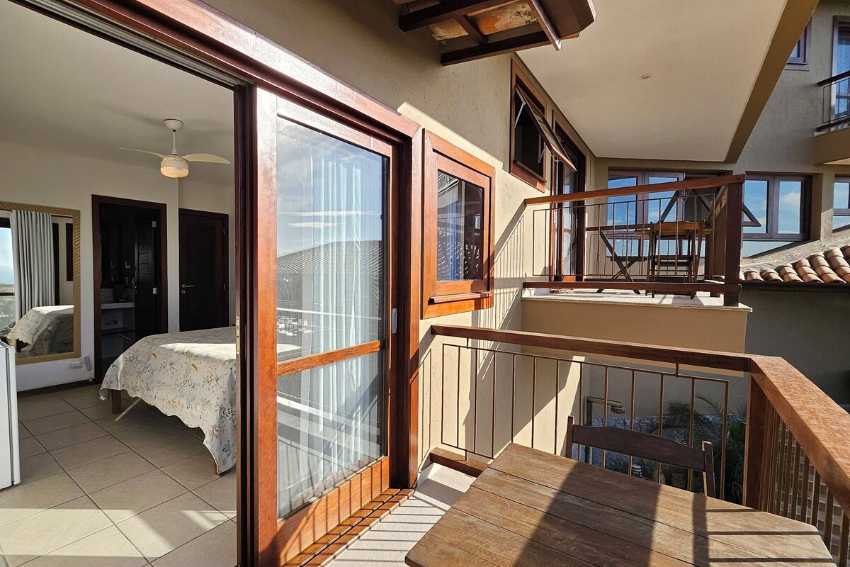 A bedroom with a bed, a desk, and a chair, a balcony with a wooden railing, and a view of the outside.