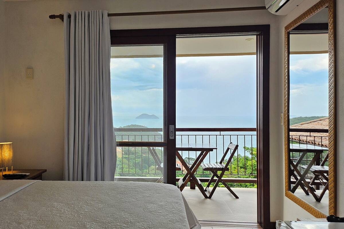 A bedroom with a bed, a nightstand, a lamp, and a view of a balcony with a table and chairs.