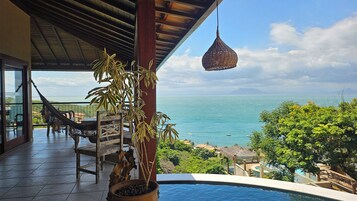 Un balcón con vista al mar, una hamaca, una silla de madera, una planta en maceta y una lámpara colgante.