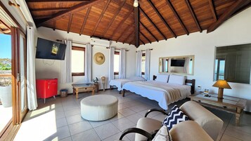Un dormitorio con una cama, televisión, un armario rojo, ventana con cortinas, un otoman redondito y una lámpara.