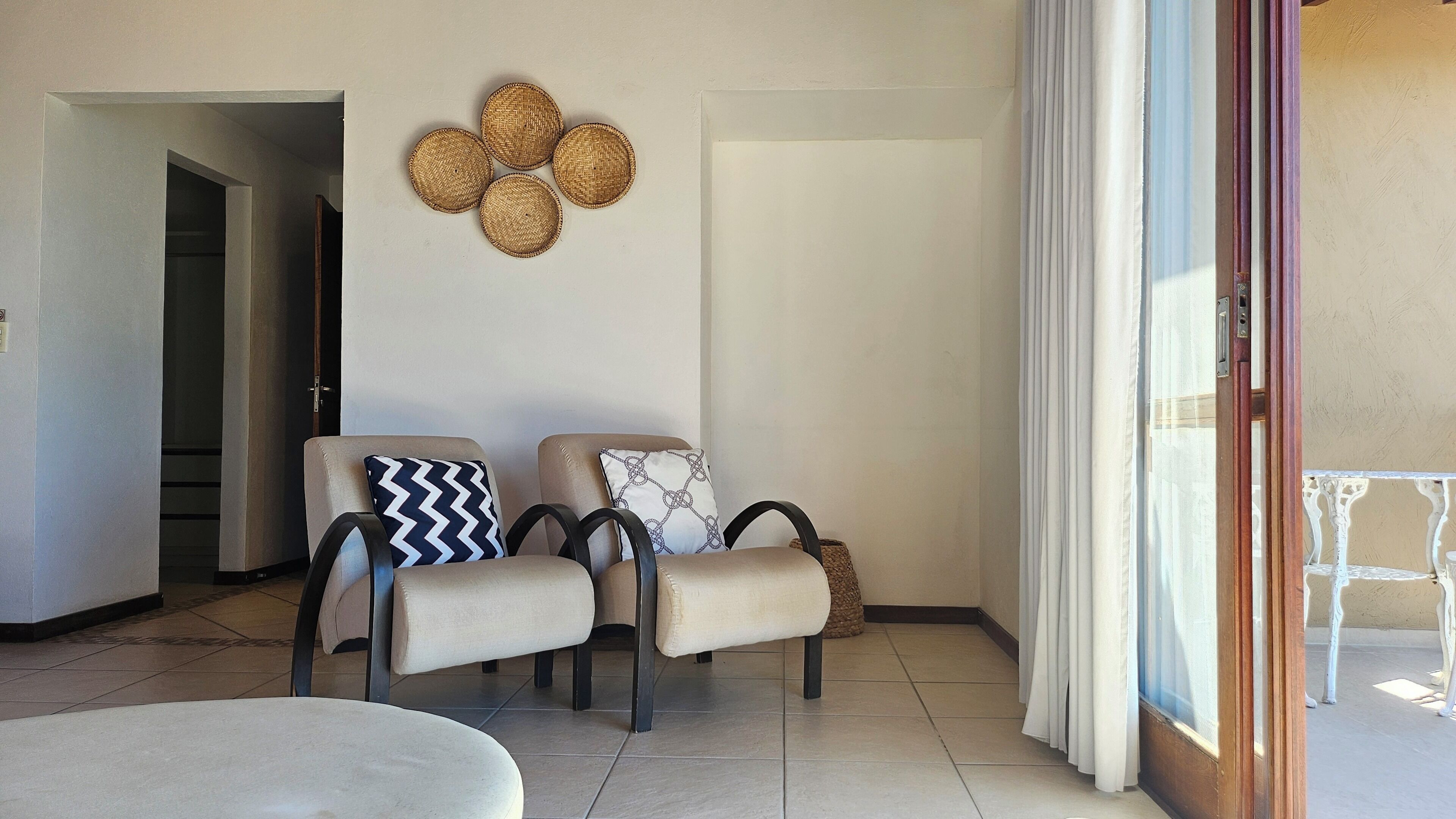 Una habitación con dos sillones, una mesita, un adorno tejido en la pared y vistas a través de una ventana con cortinas.
