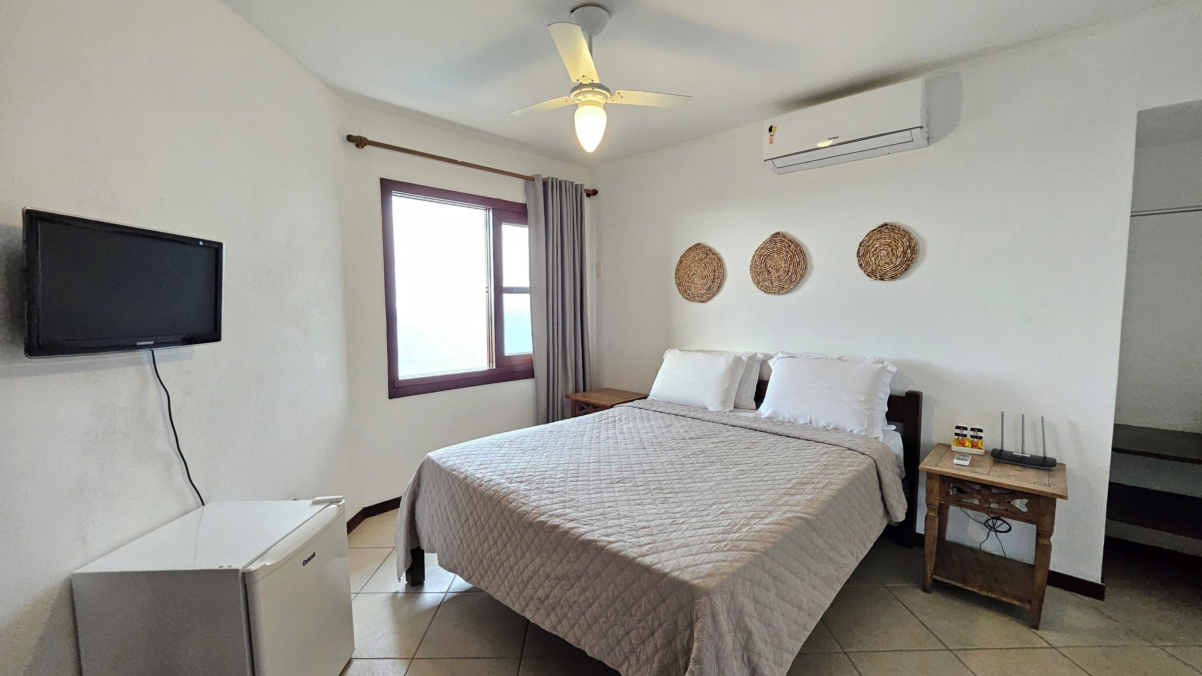 Un dormitorio con cama, televisión, aire acondicionado, ventilador de techo y ventana con cortinas.