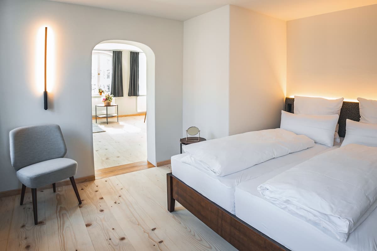 Una habitación de hotel moderna con piso de madera, un sillón gris, una cama con sábanas blancas y una mesita de noche con lámpara.