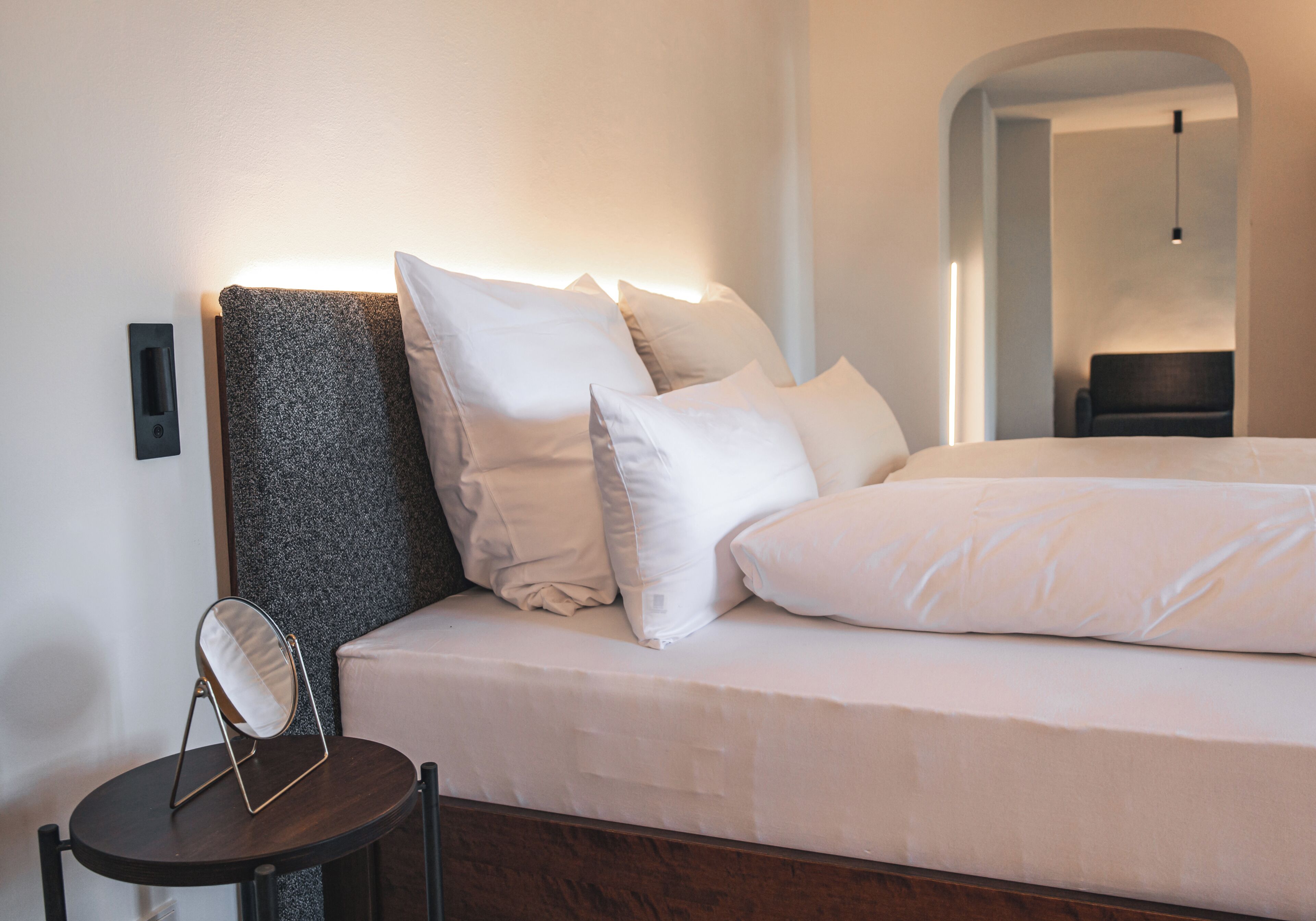Una cama bien hecha con sábanas blancas y un cabecero, una mesita redonda con un espejo y una lámpara fijada a la pared.