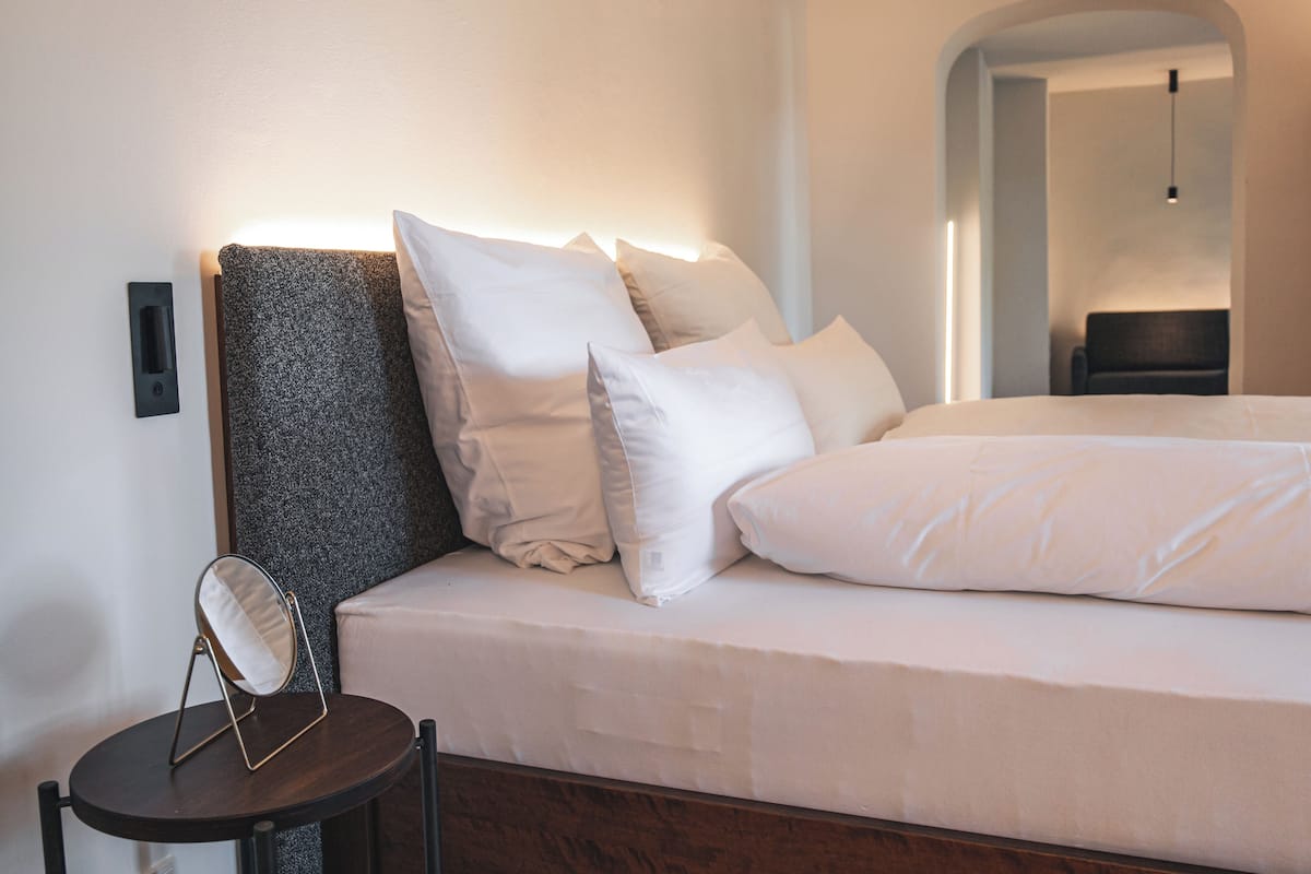 Una cama bien hecha con sábanas blancas y un cabecero, una mesita redonda con un espejo y una lámpara fijada a la pared.