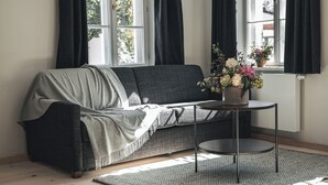 Un saló modern amb un sofà gris, una taula rodona amb un ram de flors i una finestra gran amb cortines negres.