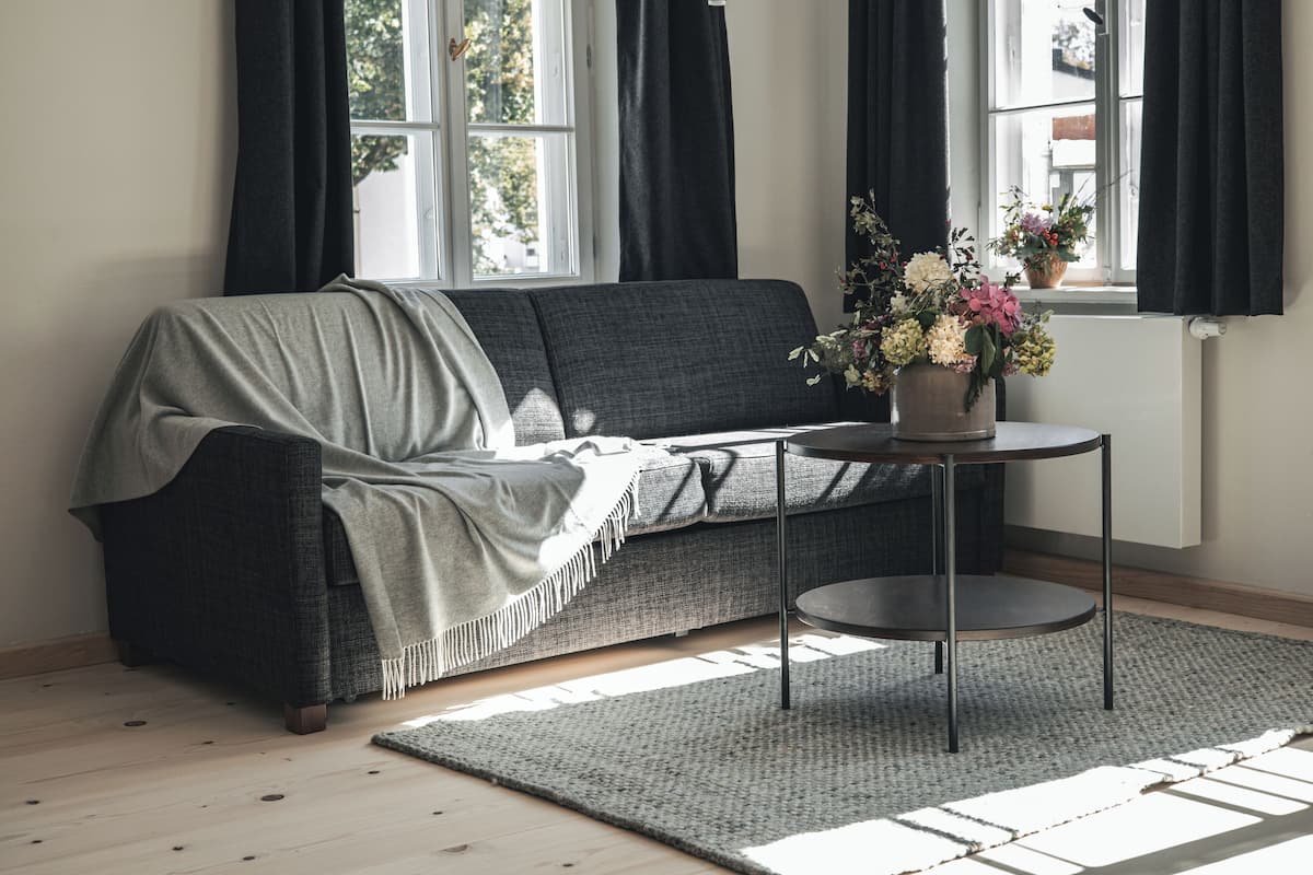 Una sala de estar moderna con un sofá gris, una mesa redonda con un jarrón de flores y un ventanal con cortinas negras.