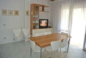 A dining area with a wooden table and white chairs, a television on a wooden cabinet, and framed pictures on the wall. - Comfortable Flat in Lignano Pineta (Lignano Sabbiadoro)