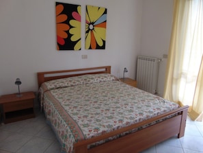 A bedroom with a wooden bed, a nightstand, a lamp, a radiator, and a window with curtains. - Cozy Apartment 150m From the sea (Lignano Sabbiadoro)