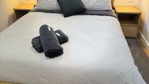 Una cama bien tendida con un edredón blanco, dos toallas oscuras y varias almohadas en una habitación de hotel.