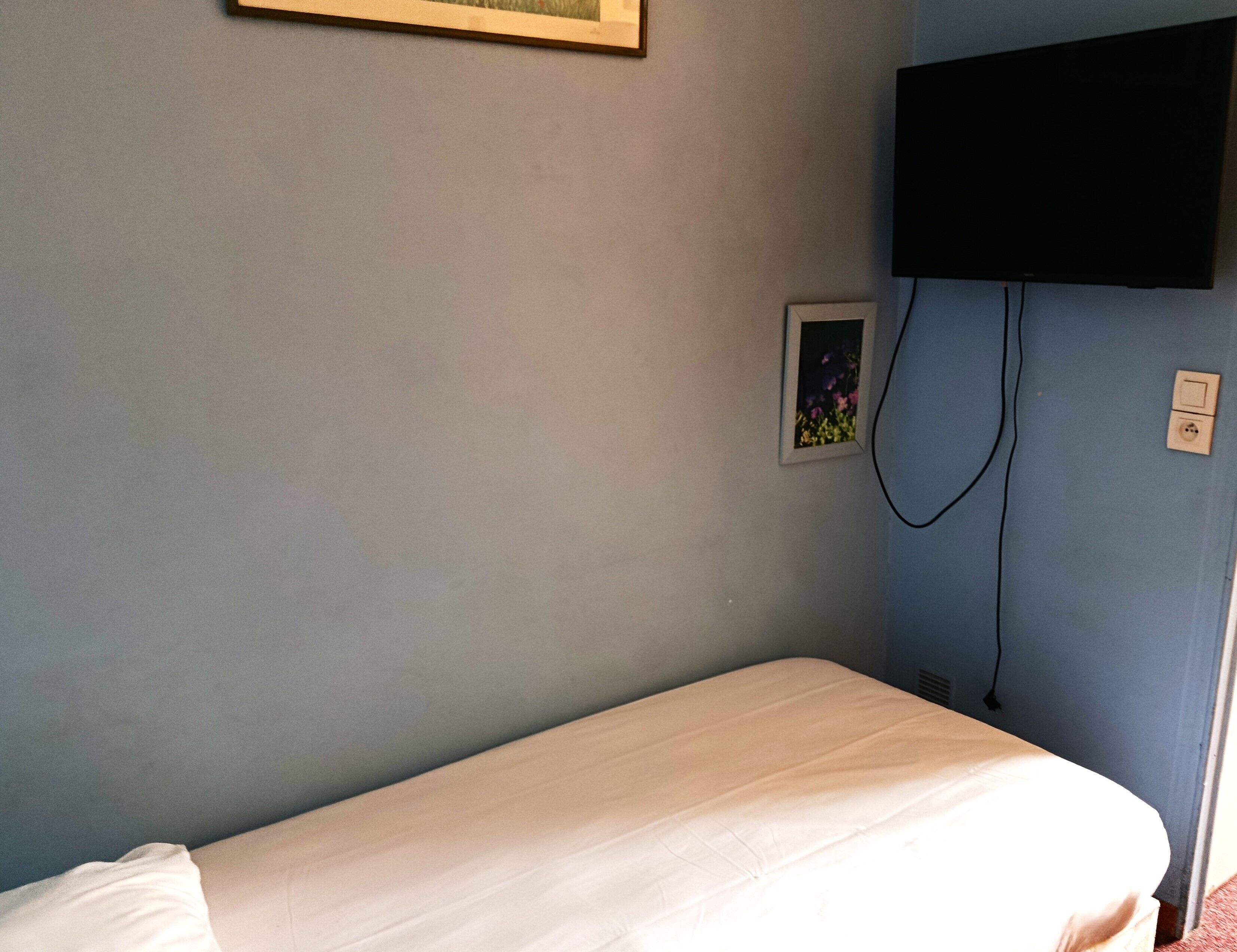 A single bed in a room with a light blue wall, a framed picture, a television mounted on the wall, and a power outlet.