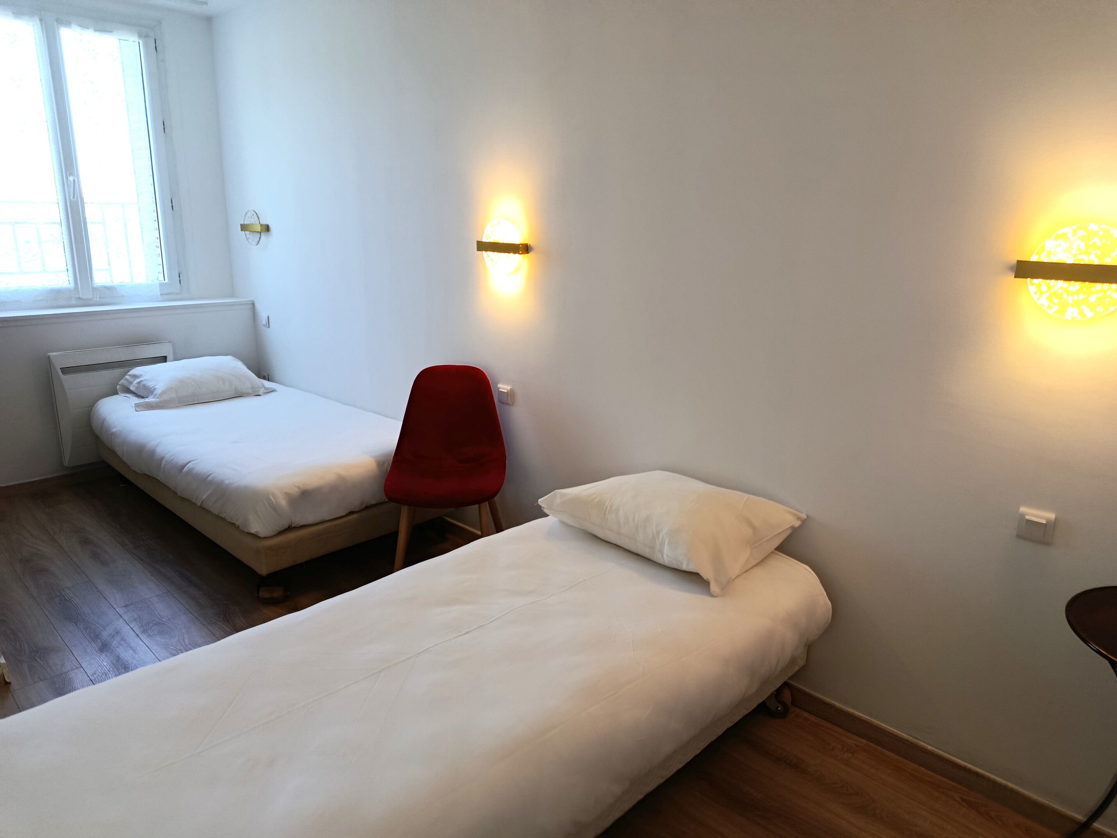 Een kamer met twee eenpersoonsbedden, een rode stoel en wandlampen.